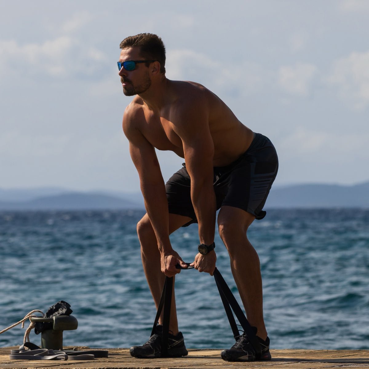 Athlete in a banded hip hinge on a seaside pier, standing on an aerobis powerband with soft knees and a long spine