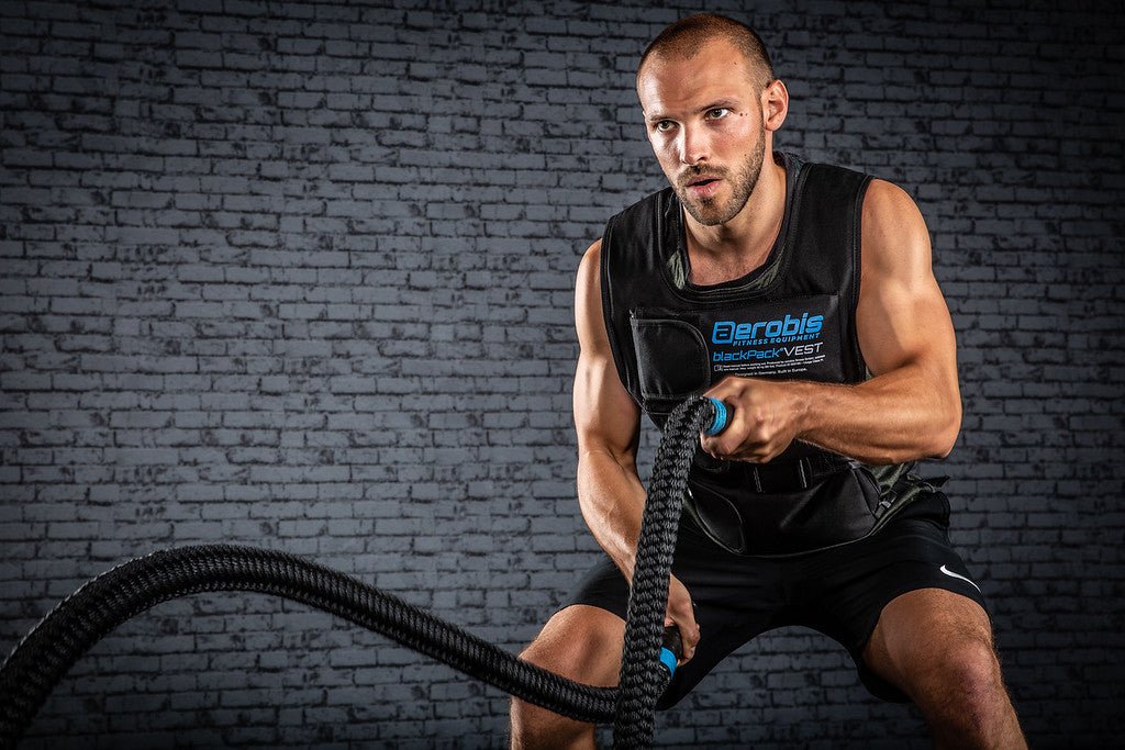 Athlete doing battle rope waves, holding both ends of a single rope against a brick wall