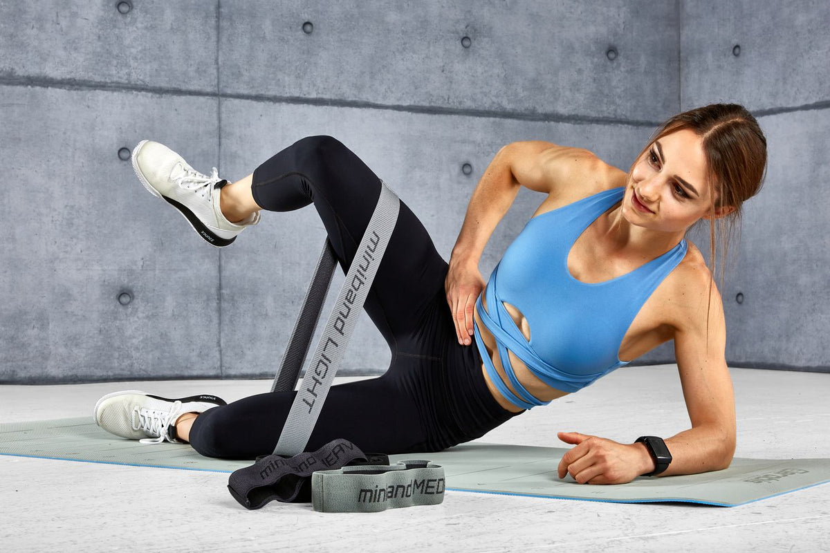 Woman lying on her side on a mat during a miniband hip exercise, propped on her forearm, top knee bent and lifted against a grey miniband around the thighs, two more minibands on the floor