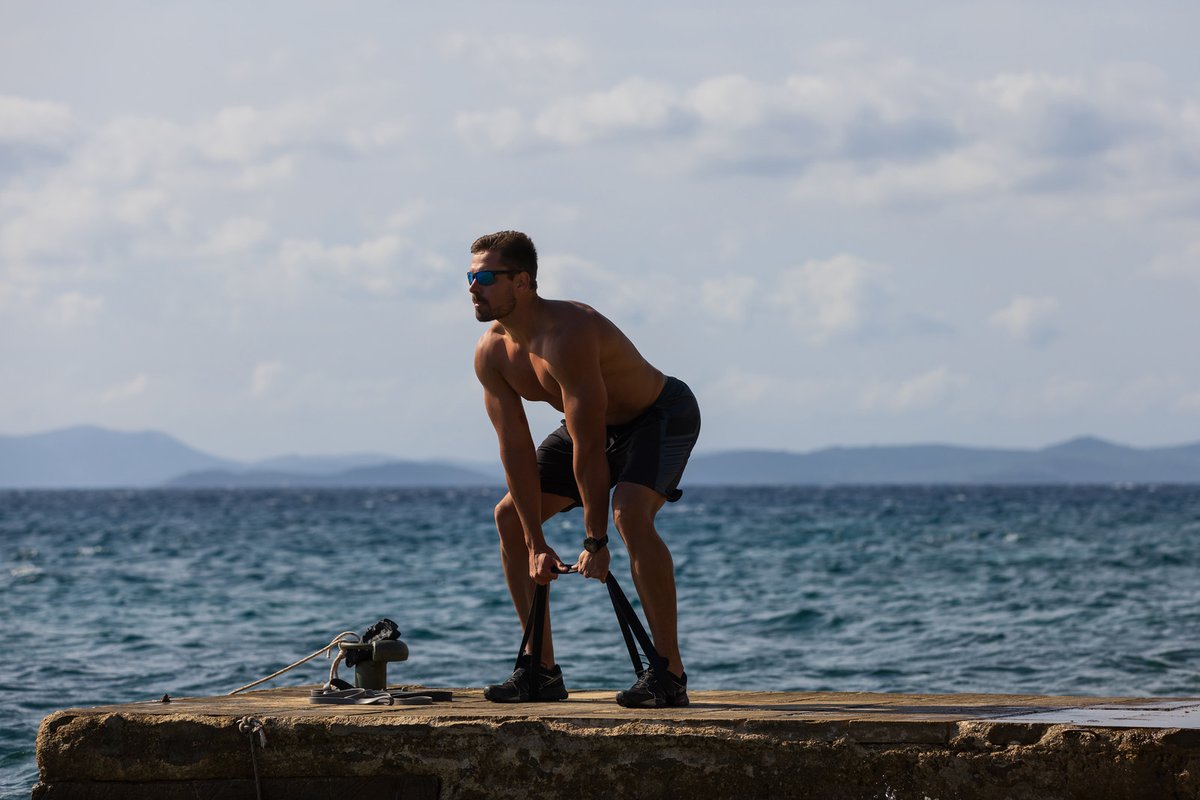 Athlete performing a resistance band Romanian deadlift, standing on an aerobis powerband in a hip hinge on a seaside pier