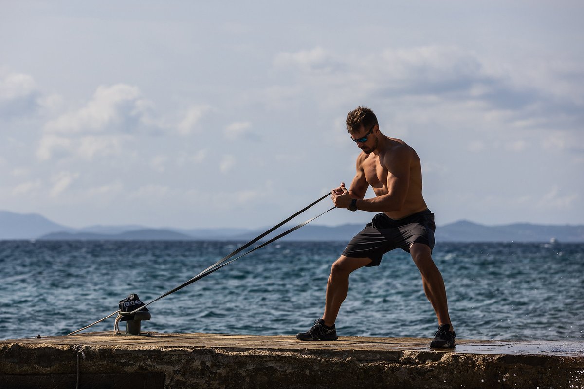 Athlete performing a standing resistance-band row with an aerobis powerband anchored on a seaside pier