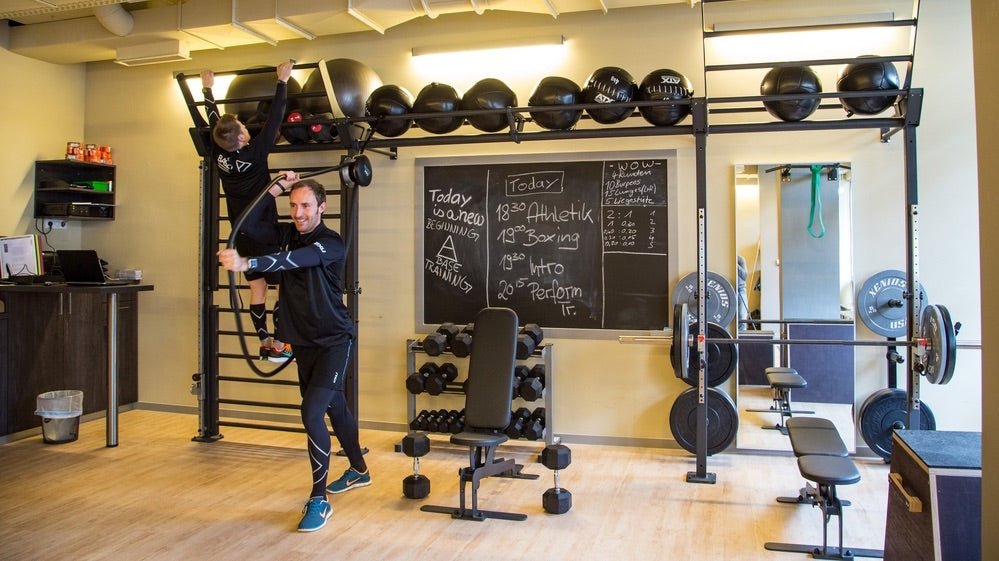 Two men exercising in a gym with wall bars and weights.