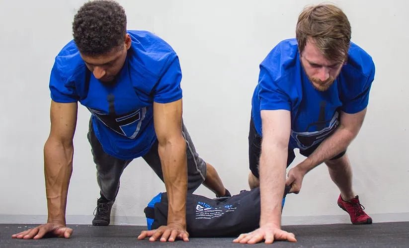 Dois homens treinam na posição de prancha com sandbag para obterem força funcional.